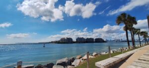 Panoramiczny widok na nadmorski deptak w słoneczny dzień. Na pierwszym planie widać kamienny brzeg z dużych, jasnoszarych głazów i chodnik z niskim murkiem i linowym ogrodzeniem. Po prawej stronie rośnie kilka palm, których liście porusza wiatr. Po lewej stronie rozciąga się spokojna, błękitna woda zatoki lub kanału. Na horyzoncie widać kilka nowoczesnych budynków mieszkalnych z balkonami i jasnymi elewacjami, a jeszcze dalej – zarys wieżowców dużego miasta. Niebo jest intensywnie niebieskie z białymi, rozproszonymi chmurami. Atmosfera wakacyjna i spokojna.