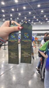 Zbliżenie dłoni trzymającej dwa zielone wstążkowe medale z napisem „FIRST Global Challenge 2025 – Panama City”. Na jednej wstążce znajduje się napis „Social Media Challenge Award”, a na drugiej „Video Storytelling Award”. Oba mają ozdobne logo wydarzenia w kolorach niebieskim, żółtym i pomarańczowym. Tło przedstawia dużą halę wystawową, w której widać uczestników ubranych w kolorowe koszulki i stanowiska z dekoracjami. Oświetlenie sztuczne, chłodne, podkreśla atmosferę zawodów i sukcesu.