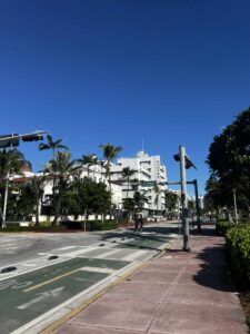 Szeroka ulica w mieście tropikalnym, z pasem rowerowym i chodnikiem po prawej stronie. Po lewej stronie rosną palmy i stoją białe budynki w stylu modernistycznym z balkonami i płaskimi dachami. Niebo jest intensywnie niebieskie, bez chmur. W oddali widać kilka osób spacerujących i jadących na rowerach.