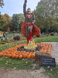 Zdjęcie wykonane w parku jesienią, przedstawiające dużą rzeźbę w kształcie postaci przypominającej superbohatera w locie, z jedną ręką uniesioną do góry. Figura wykonana jest z dziesiątek dyni i innych warzyw ułożonych kolorystycznie.
Tułów i peleryna zrobione są z pomarańczowych i brązowych dyń, nogi z czerwonych, a podstawa z mniejszych żółtych i czarnych dyni. Na piersi postaci widnieje żółto-czerwony symbol litery „S”. Rzeźba stoi na zielonym trawniku otoczonym przez dynie ułożone w okrąg.
W tle widać alejkę z drzewami o jesiennych barwach – złotych, pomarańczowych i zielonych. Po lewej stronie znajdują się grupy ludzi spacerujących po parku. Przed rzeźbą ustawiono tabliczkę z napisem w języku niemieckim: „Kunstwerke bitte nicht berühren. Danke!”, co oznacza: „Prosimy nie dotykać dzieł sztuki. Dziękujemy!”. Scena jest barwna, wesoła i jesienna.
