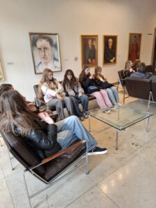 Wnętrze eleganckiego pomieszczenia, przypominającego galerię lub hol urzędu. Na ścianie wisi kilka dużych portretów mężczyzn w ramach. Na pierwszym planie grupa młodych osób, głównie dziewcząt, siedzi na brązowych skórzanych fotelach wokół szklanego stołu. Większość z nich ma długie włosy i ubrana jest w luźne, codzienne stroje – dżinsy i bluzy. Jedna z dziewczyn przegląda coś w telefonie, inna rozmawia z koleżanką.