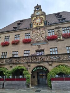 Zabytkowy budynek z piaskowcowej cegły, z ozdobnym zegarem astronomicznym na fasadzie. Zegar ma złote i czerwone elementy, kilka mniejszych tarcz i figurki. Nad wejściem do budynku widoczny jest neonowy napis „Ratskeller” w zielonym kolorze. Na balkonie stoi kilka osób, które wyglądają na zwiedzających. Budynek ma okna ozdobione czerwonymi kwiatami w donicach, a pod balkonem rosną zielone pnącza oplatające arkady.