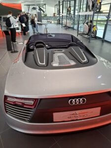 Nowoczesne wnętrze salonu lub muzeum motoryzacyjnego. Na pierwszym planie tył srebrnego samochodu sportowego marki Audi, model e-tron Spyder. Auto ma błyszczący lakier, niską sylwetkę i futurystyczne reflektory. Za samochodem widoczna jest grupa osób oglądających inne eksponaty i metalowe ramy z powieszonymi motocyklami. Przestrzeń jest duża, przeszklona, z widokiem na zewnątrz, oświetlona naturalnym światłem.