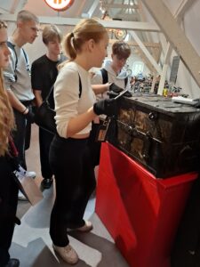 Grupa młodych osób, około sześciu, stoi wokół dużej, starej skrzyni z metalu i drewna. Skrzynia ma ciemny, brązowo-czarny kolor i widoczne metalowe okucia, zamki i zawiasy. Ustawiona jest na czerwonym podwyższeniu. Na pierwszym planie młoda kobieta w białym swetrze, czarnych legginsach i białych sportowych butach trzyma ręce na skrzyni, ma na dłoniach czarne rękawiczki ochronne. W tle widać kilka starych motocykli i eksponatów – wnętrze przypomina muzeum techniki. Światło jest ciepłe, wnętrze jasne z drewnianymi belkami pod sufitem.