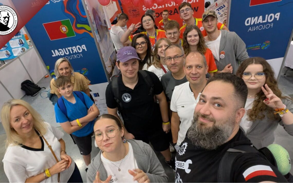 Grupa kilkunastu osób, dorosłych i młodzieży, pozuje do wspólnego zdjęcia w hali wystawowej. W tle widoczne są kolorowe ścianki z flagami Portugalii i Polski oraz napisem „Poland / Polska”. Ludzie uśmiechają się, część pokazuje znak pokoju lub unosi kciuki. Na stoiskach widać plakaty, balony i kolorowe elementy wystawowe. Scena pełna energii, przyjazna i radosna.