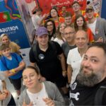 Grupa kilkunastu osób, dorosłych i młodzieży, pozuje do wspólnego zdjęcia w hali wystawowej. W tle widoczne są kolorowe ścianki z flagami Portugalii i Polski oraz napisem „Poland / Polska”. Ludzie uśmiechają się, część pokazuje znak pokoju lub unosi kciuki. Na stoiskach widać plakaty, balony i kolorowe elementy wystawowe. Scena pełna energii, przyjazna i radosna.