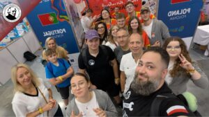 Grupa kilkunastu osób, dorosłych i młodzieży, pozuje do wspólnego zdjęcia w hali wystawowej. W tle widoczne są kolorowe ścianki z flagami Portugalii i Polski oraz napisem „Poland / Polska”. Ludzie uśmiechają się, część pokazuje znak pokoju lub unosi kciuki. Na stoiskach widać plakaty, balony i kolorowe elementy wystawowe. Scena pełna energii, przyjazna i radosna.