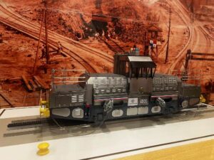 Model lokomotywy w muzeum – miniaturowy, metaliczno-szary pojazd z oznaczeniem „100” na boku, ustawiony na torach wystawowych. Lokomotywa ma druciane barierki, reflektory i detale techniczne. W tle duże zdjęcie z epoki przedstawiające budowę kanału – widoczne tory, ziemia w kolorze rdzawym i pracujący robotnicy.