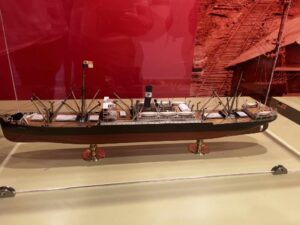 Model statku pasażerskiego lub transportowego z pierwszej połowy XX wieku, ustawiony w gablocie muzealnej. Kadłub jest czarny z brązowym pokładem i białą nadbudówką, statek ma kilka masztów z linami. Na kominie widnieje czarne oznaczenie z literą. Tło w kolorze czerwono-brązowym przedstawia sceny budowy portu i torów.