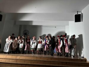 Zdjęcie przedstawia grupę około piętnastu młodych osób, stojących na drewnianej scenie w szkolnej sali. Światło jest punktowe i pada z góry, tworząc wyraźne cienie na tylnej ścianie. Tło jest jednolite, jasnoszare. Na ścianie w głębi widnieje okrągłe logo szkoły: czarna sylwetka mężczyzny w profilu na białym tle, wokół napis „I Liceum Ogólnokształcące im. S. Staszica w Lublinie”. Wszyscy uczniowie pochylają się lekko do przodu, jakby kończyli występ i kłaniali się publiczności. Ustawienie i stroje Od lewej do prawej: Kilka dziewcząt w jasnych, prostych strojach ludowych – białe lub kremowe sukienki, bufiaste rękawy, wiązania pod szyją. Część z nich ma zaplecione warkocze. Buty: czarne kozaki lub trzewiki. Następnie chłopcy i dziewczęta w tradycyjnych strojach krakowskich: Dziewczęta mają kolorowe, kwieciste wianki, czerwone korale, białe koszule i intensywnie zdobione gorsety – czerwone, czarne lub zielone, haftowane kwiaty. Spódnice wielowarstwowe, sięgające łydek – dominują kolory zielony i czerwony. Chłopcy mają na sobie: białe koszule, czerwone pasy, granatowe lub niebieskie kamizelki z naszywanymi tasiemkami i cekinami, pasiaki (luźne spodnie w czerwono-białe pionowe pasy) oraz wysokie czarne buty. Na głowach charakterystyczne czerwone czapki krakowskie z piórami – głównie pawimi. W prawej części zdjęcia stoją uczniowie w prostszych kostiumach – mężczyzna w czarnym stroju, kilka osób w białych koszulach, jedna osoba podpiera się długim kijem. Wszyscy stoją blisko siebie, niektórzy lekko śmieją się, inni zachowują powagę charakterystyczną dla finałowego ukłonu w przedstawieniu.