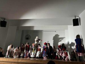 Ta fotografia ma bardziej teatralny, dramatyczny klimat. Oświetlenie znowu punktowe, mocno rzuca cienie na ścianę, w tym bardzo wyraźny cień skrzypka z uniesionym smyczkiem. Na środku stoi osoba przebrana za wiejskiego grajka: Ubrana w długą, kremową, grubą koszulę lub kaftan. Ma na sobie białą lub kremową chustę narzuconą na głowę i twarz, owijającą policzki i szyję, przez co rysy są niemal niewidoczne. Na głowie szeroki, słomkowy kapelusz. Gra na skrzypcach – instrument ma jasnobrązowy korpus, jest dobrze oświetlony, kontrastuje z jasnym strojem. Wokół skrzypka – przed nim i po jego bokach – większość pozostałych osób klęczy lub pochyla głowy. Kolory strojów takie jak w zdjęciu pierwszym – stroje krakowskie i proste stroje ludowe. Po prawej stronie zdjęcia jedna osoba stoi tyłem lub bokiem, przy ścianie. Trzyma kij i jest częściowo w cieniu. Atmosfera sceny sugeruje moment patetyczny lub symboliczny w spektaklu, być może nawiązujący do tradycji ludowych lub obrzędów.