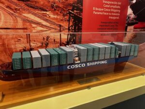 Model dużego statku kontenerowego w muzeum, o nazwie „Cosco Shipping”. Kadłub w kolorze granatowym, z białym nadpisem i licznymi zielonymi kontenerami ustawionymi w rzędach na pokładzie. W tle plansza z czerwonym napisem w języku hiszpańskim informująca o inauguracji rozbudowanego Kanału Panamskiego.
