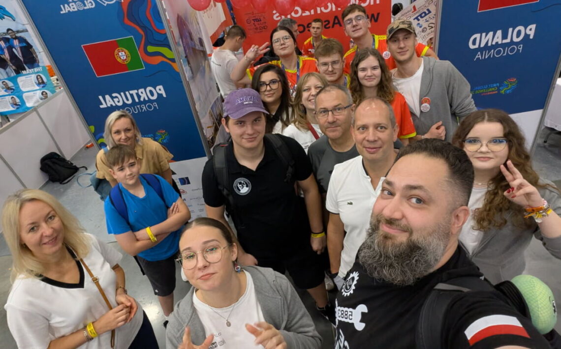 Grupa kilkunastu osób, dorosłych i młodzieży, pozuje do wspólnego zdjęcia w hali wystawowej. W tle widoczne są kolorowe ścianki z flagami Portugalii i Polski oraz napisem „Poland / Polska”. Ludzie uśmiechają się, część pokazuje znak pokoju lub unosi kciuki. Na stoiskach widać plakaty, balony i kolorowe elementy wystawowe. Scena pełna energii, przyjazna i radosna.