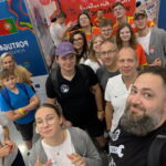 Grupa kilkunastu osób, dorosłych i młodzieży, pozuje do wspólnego zdjęcia w hali wystawowej. W tle widoczne są kolorowe ścianki z flagami Portugalii i Polski oraz napisem „Poland / Polska”. Ludzie uśmiechają się, część pokazuje znak pokoju lub unosi kciuki. Na stoiskach widać plakaty, balony i kolorowe elementy wystawowe. Scena pełna energii, przyjazna i radosna.