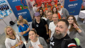 Grupa kilkunastu osób, dorosłych i młodzieży, pozuje do wspólnego zdjęcia w hali wystawowej. W tle widoczne są kolorowe ścianki z flagami Portugalii i Polski oraz napisem „Poland / Polska”. Ludzie uśmiechają się, część pokazuje znak pokoju lub unosi kciuki. Na stoiskach widać plakaty, balony i kolorowe elementy wystawowe. Scena pełna energii, przyjazna i radosna.