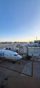 Widok z lotniska na stojący przy rękawie pasażerskim samolot linii „Viva Aerobus” (częściowo widoczne napisy). Samolot jest biały z fioletowym ogonem. W tle rozległa płyta lotniska z innymi samolotami, budynkami i błękitnym niebem o świcie lub zachodzie – słońce nisko nad horyzontem oświetla scenę miękkim światłem.