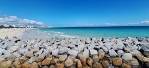 Panoramiczny widok plaży i oceanu. Na pierwszym planie duże, szare i brązowe głazy tworzące falochron. Dalej biały piasek, błękitne morze i kilka osób kąpiących się w wodzie lub spacerujących po plaży. Po lewej stronie w oddali widać miasto z białymi budynkami. Niebo czyste, lazurowe.