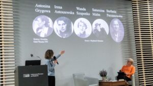 Na pierwszym zdjęciu widoczna jest scena w sali konferencyjnej lub muzealnej. Po lewej stronie stoi kobieta z krótkimi, kręconymi włosami, ubrana w jasnoniebieską marynarkę. Trzyma mikrofon w prawej ręce i lewą dłonią wskazuje na dużą projekcję na ścianie za sobą. Po prawej stronie, w fotelu, siedzi druga kobieta ubrana w pomarańczową bluzkę i czarne spodnie; trzyma notes lub teczkę na kolanach i spogląda na prelegentkę.
Na ekranie widać pięć czarno-białych zdjęć portretowych kobiet w okrągłych ramkach. Nad zdjęciami widnieją podpisy: „Antonina Grygowa”, „Irena Antoszewska”, „Wanda Szupenko”, „Saturnina Malm” i „Stefania Perzanowska”. Pod zdjęciami znajdują się napisy w języku angielskim informujące o źródłach: „Source: Grodzka Gate/Theater NN” oraz „Source: Majdanek Museum”. Tło projekcji jest szare. Na pierwszym planie stoi czarna mównica z napisem: „Państwowe Muzeum na Majdanku” oraz logo składające się z kilku poziomych linii.