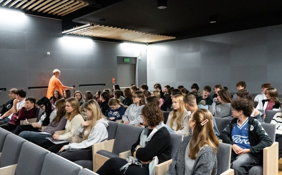 Na drugim zdjęciu widać wnętrze sali audytoryjnej z rzędami nowoczesnych, szarych foteli. Sala jest dobrze oświetlona, ściany i sufit w kolorze szarym, z panelami akustycznymi i drewnianymi listwami. W ławkach siedzi kilkudziesięciu młodych ludzi — uczniów lub studentów, głównie w wieku nastoletnim. Słuchają uważnie, niektórzy notują. W tle, po lewej stronie, kobieta w pomarańczowej bluzce (ta sama co z pierwszego zdjęcia) przechodzi między rzędami z mikrofonem w ręce. Wnętrze ma nowoczesny, minimalistyczny styl.