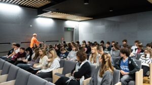 Na drugim zdjęciu widać wnętrze sali audytoryjnej z rzędami nowoczesnych, szarych foteli. Sala jest dobrze oświetlona, ściany i sufit w kolorze szarym, z panelami akustycznymi i drewnianymi listwami. W ławkach siedzi kilkudziesięciu młodych ludzi — uczniów lub studentów, głównie w wieku nastoletnim. Słuchają uważnie, niektórzy notują. W tle, po lewej stronie, kobieta w pomarańczowej bluzce (ta sama co z pierwszego zdjęcia) przechodzi między rzędami z mikrofonem w ręce. Wnętrze ma nowoczesny, minimalistyczny styl.