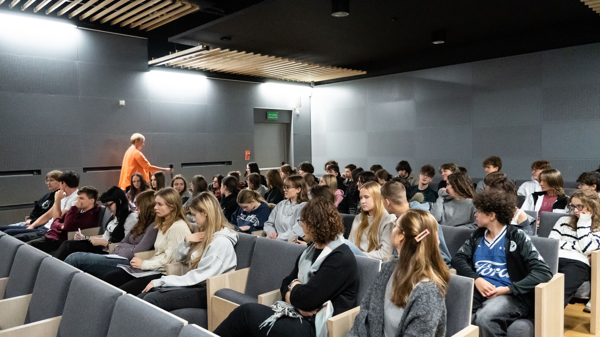 Na drugim zdjęciu widać wnętrze sali audytoryjnej z rzędami nowoczesnych, szarych foteli. Sala jest dobrze oświetlona, ściany i sufit w kolorze szarym, z panelami akustycznymi i drewnianymi listwami. W ławkach siedzi kilkudziesięciu młodych ludzi — uczniów lub studentów, głównie w wieku nastoletnim. Słuchają uważnie, niektórzy notują. W tle, po lewej stronie, kobieta w pomarańczowej bluzce (ta sama co z pierwszego zdjęcia) przechodzi między rzędami z mikrofonem w ręce. Wnętrze ma nowoczesny, minimalistyczny styl.