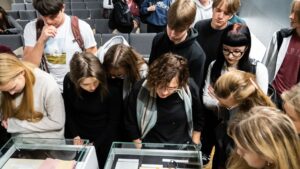 Grupa młodych osób stoi nad szklanymi gablotami muzealnymi. W gablotach leżą dokumenty i przedmioty, prawdopodobnie pamiątki historyczne — widać kartki papieru, koperty, metalowy pierścień i bransoletkę. Uczniowie pochylają się, uważnie oglądając eksponaty. Większość z nich ma poważne, skupione twarze. Wszyscy ubrani są w codzienne ubrania: bluzy, swetry, ciemne kurtki. Tło stanowią rzędy szarych foteli, takie jak w sali konferencyjnej.