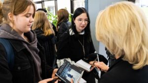 Trzy młode kobiety stoją we wnętrzu budynku, prawdopodobnie w przedsionku muzeum. Na pierwszym planie jedna z nich, o jasnych włosach, trzyma książkę z tytułem „Fałszywa hrabina” (autorki Elizabeth B. White i Joanny Sliwy). Książka ma ciemną okładkę z niebieskim napisem i wizerunkiem sylwetki kobiety idącej po zaśnieżonej ulicy. Kobieta podaje książkę starszej blondynce, która trzyma długopis, prawdopodobnie w celu uzyskania autografu. Obok młoda kobieta z ciemnymi włosami i okrągłymi okularami rozmawia z autorką. W tle widać inne osoby i zielone rośliny doniczkowe.
