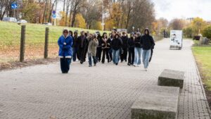 Na ostatnim zdjęciu widać grupę około dwudziestu młodych osób idących po szerokim, brukowanym chodniku w terenie otwartym. Na czele grupy idzie przewodniczka ubrana w niebieską, grubą kurtkę z kapturem obszytym futrem, na szyi ma identyfikator. Uczestnicy wycieczki są ubrani w ciepłe kurtki, bluzy z kapturem i czapki — sugeruje to chłodną pogodę, prawdopodobnie jesienną. W tle po lewej stronie widoczny jest trawnik z drzewami o żółtych i pomarańczowych liściach, a także ogrodzenie z drutu kolczastego na betonowych słupkach. W głębi kadru stoi tablica lub ekspozycja plenerowa z napisem „Sobibór”.