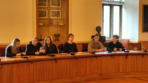 Siedem młodych osób, trzy dziewczyny i czterech chłopców, siedzi przy długim, drewnianym stole w dużej sali posiedzeń. Na stole ustawione są mikrofony. W tle widać okno z kratą i półki z pamiątkami oraz popiersiem z brązu. Ściany są kremowe, wystrój klasyczny. Uczniowie mają notatniki i długopisy, wyglądają, jakby uczestniczyli w spotkaniu lub warsztacie.