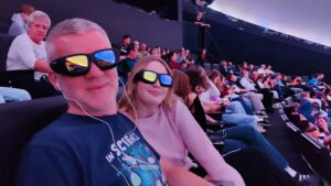 Duża sala kinowa lub planetarium. Na pierwszym planie mężczyzna i młoda kobieta siedzą obok siebie w ciemnych fotelach. Mają na uszach słuchawki i na oczach duże okulary 3D z odbiciem kolorowych świateł. Za nimi widać wielu innych widzów w podobnych okularach. Wnętrze jest półciemne, z niebiesko-fioletowym oświetleniem, co tworzy futurystyczny klimat.