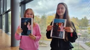 Dwie dziewczyny stoją obok siebie w jasnym korytarzu z dużymi oknami. W rękach trzymają wydrukowane zdjęcia. Dziewczyna po lewej, w różowej bluzie, pokazuje zdjęcie dwóch osób na tle kosmosu – z gwiazdami i planetami. Dziewczyna po prawej, w czarnej bluzie, trzyma podobne zdjęcie, ale z tłem przedstawiającym wybuch wulkanu z lawą. Obie uśmiechają się, a za nimi widać drzewa z jesiennymi, żółtymi liśćmi.