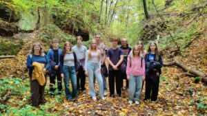 Grupa około dwunastu osób stoi w lesie. Drzewa mają żółto-zielone liście, na ziemi leży dużo jesiennych liści. Ludzie ustawieni są w dwóch rzędach, patrzą w stronę aparatu. Większość z nich to młodzież, z przodu dwie dorosłe osoby – kobieta po lewej z uśmiechem trzyma żółtą kurtkę. Ścieżka w lesie jest wąska, otoczona skałami i krzewami.