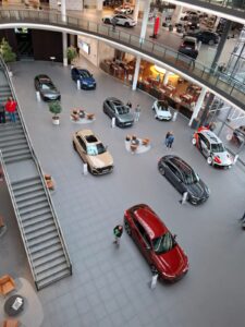 Wnętrze dużego, nowoczesnego salonu samochodowego lub muzeum motoryzacji, oglądane z góry z antresoli. Na szerokiej, szarej posadzce ustawionych jest dziewięć samochodów różnych modeli marki Audi. Auta mają różne kolory – wśród nich wyróżnia się czerwony samochód na pierwszym planie, beżowy w centralnej części oraz niebieski i szary dalej w tle. Samochody są rozmieszczone w równych odstępach, każdy z tabliczką informacyjną obok.
Po lewej stronie zdjęcia widoczne są szerokie schody prowadzące na górny poziom z balustradą ze szkła. Wokół samochodów stoją nowoczesne stoliki i brązowe fotele na metalowych nogach. W tle, za przeszkloną ścianą, znajduje się restauracja z pomarańczowym oświetleniem i kilkoma stolikami. Na górnym poziomie, widocznym w tle, stoją kolejne samochody wystawowe. Kilka osób spaceruje po hali i ogląda eksponaty. Wnętrze jest jasne, przestronne i eleganckie, z dominującą szarą i srebrną kolorystyką.