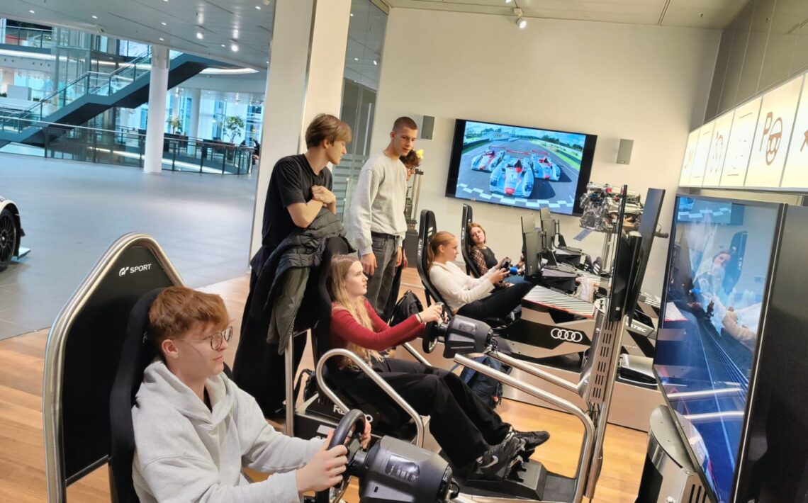 Grupa młodych osób w nowoczesnym wnętrzu z samochodami sportowymi. Część z nich siedzi w fotelach symulatorów jazdy – trzymają kierownice i patrzą w duże ekrany, na których widać tor wyścigowy i samochody. Pozostali stoją obok i obserwują. W tle widać przeszklone piętro i wystawione samochody wyścigowe. Pomieszczenie jest jasne, nowoczesne i przestronne.