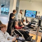 Grupa młodych osób w nowoczesnym wnętrzu z samochodami sportowymi. Część z nich siedzi w fotelach symulatorów jazdy – trzymają kierownice i patrzą w duże ekrany, na których widać tor wyścigowy i samochody. Pozostali stoją obok i obserwują. W tle widać przeszklone piętro i wystawione samochody wyścigowe. Pomieszczenie jest jasne, nowoczesne i przestronne.