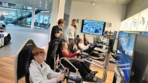 Grupa młodych osób w nowoczesnym wnętrzu z samochodami sportowymi. Część z nich siedzi w fotelach symulatorów jazdy – trzymają kierownice i patrzą w duże ekrany, na których widać tor wyścigowy i samochody. Pozostali stoją obok i obserwują. W tle widać przeszklone piętro i wystawione samochody wyścigowe. Pomieszczenie jest jasne, nowoczesne i przestronne.