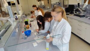 Laboratorium naukowe. Kilka dziewcząt w białych fartuchach i okularach ochronnych pracuje przy długim stole laboratoryjnym. Na stole stoją naczynia, butelki, pipety, miski i kartki z instrukcjami. Uczennice skupione są na wykonywaniu doświadczenia – jedna miesza substancje, inna nalewa płyn z kartonowego opakowania. W tle widać komputery, zlewki i sprzęt laboratoryjny.