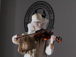 To zbliżenie na tę samą postać, która grała na skrzypcach. Wygląd postaci Osoba jest cała ubrana w jasne tkaniny – beżowe, kremowe, białe. Twarz zakryta delikatnym woalem z cienkiego materiału, który opada z kapelusza i sprawia, że rysy twarzy są niemal niewidoczne. Na ramieniu ma narzucony sweter lub narzutę z grubym splotem, w naturalnych kolorach. W rękach trzyma skrzypce w ciepłym, brązowym kolorze. Skrzypce są dobrze oświetlone, przez co lakier instrumentu błyszczy. W tle widać fragment szkolnego logo.