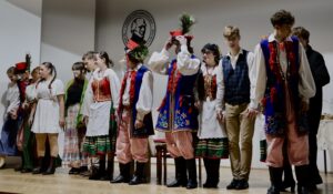 Zdjęcie przedstawia całą grupę uczniów stojących w szeregu, tym razem w jaśniejszym, równomiernym świetle, co pozwala dobrze zobaczyć detale strojów. Stroje i ustawienie Od lewej do prawej widzimy: Uczniów w jasnych, prostych sukienkach lub koszulach, z warkoczami, naturalnymi włosami, bez nakryć głowy. Dziewczęta w strojach krakowskich – bogato zdobione gorsety, kolorowe spódnice, czerwone korale. Chłopców w tradycyjnych strojach krakowskich: czerwone czapki z pawimi piórami, pasiaki, granatowe kamizelki ozdobione czerwonymi chwostami i haftami kwiatowymi. Pojawia się też chłopak w jasnej koszuli i kamizelce w granatową kratę. Wszyscy stoją spokojnie, część trzyma się za ręce. Twarze mają naturalne, z lekkimi uśmiechami.