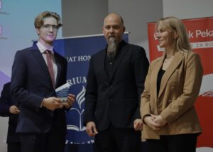 Na zdjęciu znajdują się trzy osoby stojące na tle roll-upów. Od lewej: młody chłopak w granatowym garniturze, białej koszuli i bordowym krawacie — trzyma statuetkę nagrody, dumnie prezentując ją przed sobą. Ma jasne włosy, ułożone w fale, oraz okulary.

Obok niego stoi mężczyzna o charakterystycznej, długiej, gęstej brodzie sięgającej klatki piersiowej. Ubrany jest całkowicie na czarno: czarna marynarka, koszula, spodnie. Patrzy spokojnie przed siebie.

Po prawej stronie stoi kobieta w jasnobrązowej, eleganckiej marynarce. Ma blond włosy do ramion. Uśmiecha się delikatnie i trzyma dłonie splecione przed sobą.

Za nimi widoczne są roll-upy Kuratorium Oświaty (granatowy) oraz Banku Pekao (czerwony).