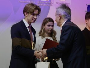 Zbliżenie na moment wręczania nagrody. Młody chłopak w ciemnym garniturze i bordowym krawacie ściska dłoń starszego mężczyzny ubranego w granatowy garnitur. Chłopak trzyma białą torebkę prezentową i akrylową statuetkę nagrodową.

Za nimi stoi młoda kobieta z długimi blond włosami, ubrana w białą bluzkę. W dłoni trzyma czerwoną teczkę. Patrzy w stronę mężczyzny wręczającego nagrodę.

Tło jest rozświetlone fioletowym odcieniem pochodzącym z rzutnika komputerowego.