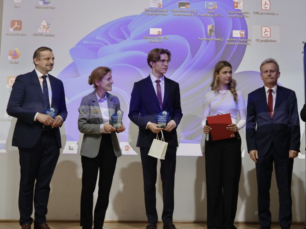 Na scenie stoi pięć osób, ustawionych w szeregu. Tło stanowi duży ekran projektora, na którym widać pulpit komputera z ikonami programów i fioletową grafiką systemową. Od lewej: Mężczyzna w granatowym garniturze i niebieskim krawacie, uśmiecha się. Trzyma w rękach małą szklaną lub akrylową nagrodę w kształcie trofeum. Kobieta w szarej marynarce i jasno-fioletowej koszuli. Trzyma podobną nagrodę w obu dłoniach i uśmiecha się, patrząc na stojącego obok niej młodzieńca. Młody chłopak w eleganckim garniturze, białej koszuli i bordowym krawacie. Ma jasne, dłuższe włosy i okulary. Trzyma nagrodę oraz jasną papierową torebkę. Młoda kobieta w białej bluzce i czarnych spodniach. Długie blond włosy opadają jej na ramiona. Trzyma czerwoną teczkę i długopis. Mężczyzna w ciemnym garniturze i czerwonym krawacie stoi po prawej stronie, patrząc przed siebie z neutralnym wyrazem twarzy. Zdjęcie przedstawia prawdopodobnie moment uroczystego wręczenia wyróżnień lub nagród.