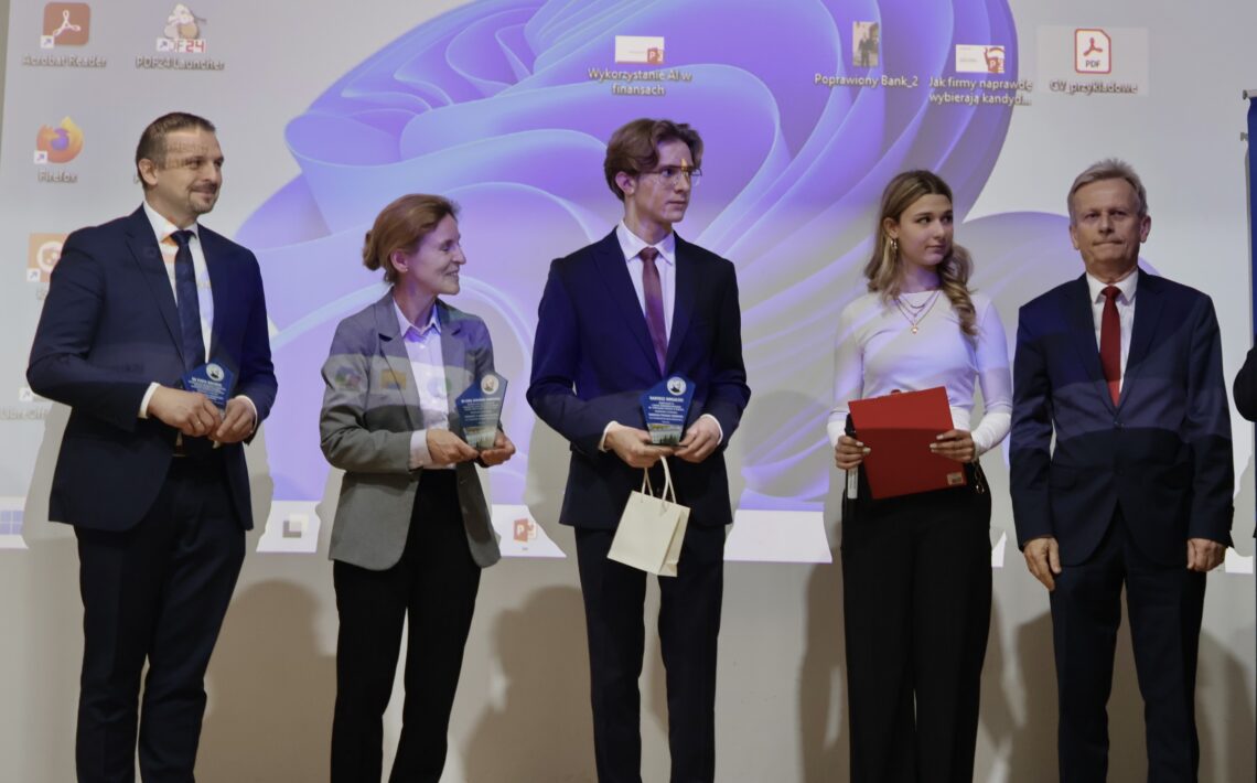 Na scenie stoi pięć osób, ustawionych w szeregu. Tło stanowi duży ekran projektora, na którym widać pulpit komputera z ikonami programów i fioletową grafiką systemową. Od lewej: Mężczyzna w granatowym garniturze i niebieskim krawacie, uśmiecha się. Trzyma w rękach małą szklaną lub akrylową nagrodę w kształcie trofeum. Kobieta w szarej marynarce i jasno-fioletowej koszuli. Trzyma podobną nagrodę w obu dłoniach i uśmiecha się, patrząc na stojącego obok niej młodzieńca. Młody chłopak w eleganckim garniturze, białej koszuli i bordowym krawacie. Ma jasne, dłuższe włosy i okulary. Trzyma nagrodę oraz jasną papierową torebkę. Młoda kobieta w białej bluzce i czarnych spodniach. Długie blond włosy opadają jej na ramiona. Trzyma czerwoną teczkę i długopis. Mężczyzna w ciemnym garniturze i czerwonym krawacie stoi po prawej stronie, patrząc przed siebie z neutralnym wyrazem twarzy. Zdjęcie przedstawia prawdopodobnie moment uroczystego wręczenia wyróżnień lub nagród.