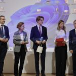 Na scenie stoi pięć osób, ustawionych w szeregu. Tło stanowi duży ekran projektora, na którym widać pulpit komputera z ikonami programów i fioletową grafiką systemową. Od lewej: Mężczyzna w granatowym garniturze i niebieskim krawacie, uśmiecha się. Trzyma w rękach małą szklaną lub akrylową nagrodę w kształcie trofeum. Kobieta w szarej marynarce i jasno-fioletowej koszuli. Trzyma podobną nagrodę w obu dłoniach i uśmiecha się, patrząc na stojącego obok niej młodzieńca. Młody chłopak w eleganckim garniturze, białej koszuli i bordowym krawacie. Ma jasne, dłuższe włosy i okulary. Trzyma nagrodę oraz jasną papierową torebkę. Młoda kobieta w białej bluzce i czarnych spodniach. Długie blond włosy opadają jej na ramiona. Trzyma czerwoną teczkę i długopis. Mężczyzna w ciemnym garniturze i czerwonym krawacie stoi po prawej stronie, patrząc przed siebie z neutralnym wyrazem twarzy. Zdjęcie przedstawia prawdopodobnie moment uroczystego wręczenia wyróżnień lub nagród.