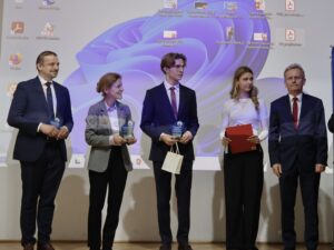 Na scenie stoi pięć osób, ustawionych w szeregu. Tło stanowi duży ekran projektora, na którym widać pulpit komputera z ikonami programów i fioletową grafiką systemową.

Od lewej:

Mężczyzna w granatowym garniturze i niebieskim krawacie, uśmiecha się. Trzyma w rękach małą szklaną lub akrylową nagrodę w kształcie trofeum.

Kobieta w szarej marynarce i jasno-fioletowej koszuli. Trzyma podobną nagrodę w obu dłoniach i uśmiecha się, patrząc na stojącego obok niej młodzieńca.

Młody chłopak w eleganckim garniturze, białej koszuli i bordowym krawacie. Ma jasne, dłuższe włosy i okulary. Trzyma nagrodę oraz jasną papierową torebkę.

Młoda kobieta w białej bluzce i czarnych spodniach. Długie blond włosy opadają jej na ramiona. Trzyma czerwoną teczkę i długopis.

Mężczyzna w ciemnym garniturze i czerwonym krawacie stoi po prawej stronie, patrząc przed siebie z neutralnym wyrazem twarzy.

Zdjęcie przedstawia prawdopodobnie moment uroczystego wręczenia wyróżnień lub nagród.