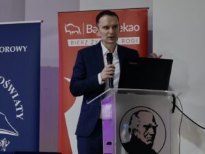 Mężczyzna w granatowym garniturze stoi za przezroczystą mównicą z czarnym wizerunkiem mężczyzny. W prawej dłoni trzyma mikrofon, a lewą wykonuje lekki gest, podkreślający jego wypowiedź. Ma krótkie, brązowe włosy, wygląda na około 30–40 lat, ma elegancki, spokojny wyraz twarzy.

Obok niego, w tle, stoi czerwony roll-up Banku Pekao z dużym białym logotypem. Po lewej stronie widoczny jest fragment granatowego roll-upu Kuratorium Oświaty.

Mężczyzna stoi lekko bokiem, widać laptopa Lenovo otwartego na mównicy. Jego styl i postawa sugerują, że prowadzi profesjonalny wykład.