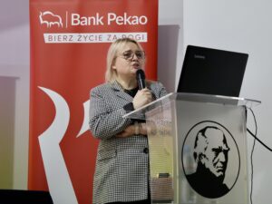 Na zdjęciu stoi kobieta przemawiająca przy przeźroczystej mównicy z czarnym nadrukiem przedstawiającym profil mężczyzny. Kobieta trzyma mikrofon w prawej dłoni. Jest ubrana w biało-czarną marynarkę w drobną pepitkę, pod którą ma czarną bluzkę. Jej blond włosy są rozpuszczone i sięgają ramion. Nosi okulary w cienkich, jasnych oprawkach.

Po lewej stronie za nią stoi duży czerwony roll-up z logotypem Banku Pekao i hasłem „Bierz życie za rogi”. Po prawej stronie pojawia się fragment białej ściany i rzutnika z otwartym laptopem marki Lenovo stojącym na mównicy.

Wyraz twarzy kobiety jest skupiony i lekko poważny, jakby przekazywała istotne informacje podczas prezentacji ekonomicznej lub finansowej.
