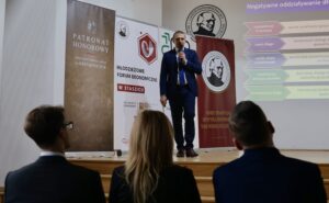 Scena z wykładu odbywającego się na szkolnej auli. Zdjęcie zrobione z perspektywy publiczności, zza pleców kilku osób siedzących w pierwszym rzędzie. Na scenie stoi mężczyzna w granatowym garniturze, białej koszuli i ciemnym krawacie. Trzyma mikrofon w prawej ręce i patrzy w stronę widowni. Jego postawa jest pewna, lekko pochylona do przodu — wygląda na osobę prowadzącą prezentację.

Za nim widoczny jest duży rzutnik wyświetlający slajd na jasnej ścianie. Slajd ma tytuł „Negatywne oddziaływanie długu” oraz zestaw kolorowych pasków z opisami — każdy pasek w innym kolorze, przedstawiający różne pojęcia ekonomiczne.

Po lewej stronie sceny stoją trzy roll-upy:

brązowy z napisem „PATRONAT HONOROWY Prezydent Miasta Lublin”,

biały z logotypami i napisem „Młodzieżowe Forum Ekonomiczne w Staszicu”,

bordowy z grafikiem profilem mężczyzny i hasłem o tradycji oraz innowacyjności.

Na pierwszym planie, rozmazane, widać sylwetki trzech słuchaczy siedzących w pierwszym rzędzie — dwóch mężczyzn i jednej kobiety.