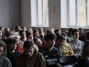 Sala szkolna wypełniona jest dużą grupą uczniów siedzących w rzędach czarnych składanych krzeseł. Kadr obejmuje ich od przodu, na wysokości twarzy i ramion. Przeważają młodzi ludzie w wieku nastoletnim — chłopcy i dziewczęta.

Światło wpada do pomieszczenia przez duże okna po prawej stronie kadru, dając jasne, naturalne oświetlenie. Uczniowie siedzą spokojnie, większość patrzy przed siebie, niektórzy w dół lub na boki.

Po lewej stronie są bardziej skupione twarze:

chłopak z ciemniejszymi, kręconymi włosami w czarnej bluzie, patrzący poważnie przed siebie;

obok chłopak w brązowej bluzie z kapturem, pochylony, jakby coś analizował;

w kolejnych rzędach widać dziewczęta i chłopców, część z okularami, część z dłuższymi włosami.

W centrum kadru, w tylnej części sali, jedna z dziewczyn opiera głowę lekko o ramię koleżanki, wygląda na zmęczoną lub znudzoną. Po prawej stronie znajduje się grupa chłopców, część ubrana w jasne bluzy — jeden w szarej, drugi w żółtej z zielonym nadrukiem.

Atmosfera jest spokojna, niektórzy wyglądają na zmęczonych, inni lekko zaciekawionych, ale całość sprawia wrażenie zwyczajnego szkolnego wykładu lub prelekcji, podczas którego młodzież słucha.