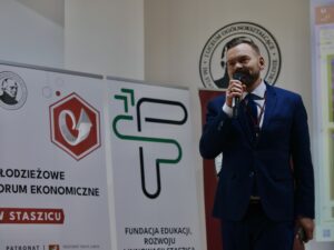 Mężczyzna w garniturze – prawdopodobnie prowadzący lub organizator – stoi z mikrofonem w prawej dłoni. Ma krótko ostrzyżone włosy i brodę. Ubrany jest w granatowy garnitur, białą koszulę i bordowy identyfikator na szyi.

Tło zdjęcia stanowią trzy roll-upy: biały z czerwonym logotypem przypominającym strzałkę lub symbol ekonomiczny, biały z zielonym logo fundacji oraz bordowy przedstawiający herb lub portret mężczyzny. Mężczyzna wygląda na osobę prowadzącą prezentację lub przemówienie podczas wydarzenia edukacyjnego.